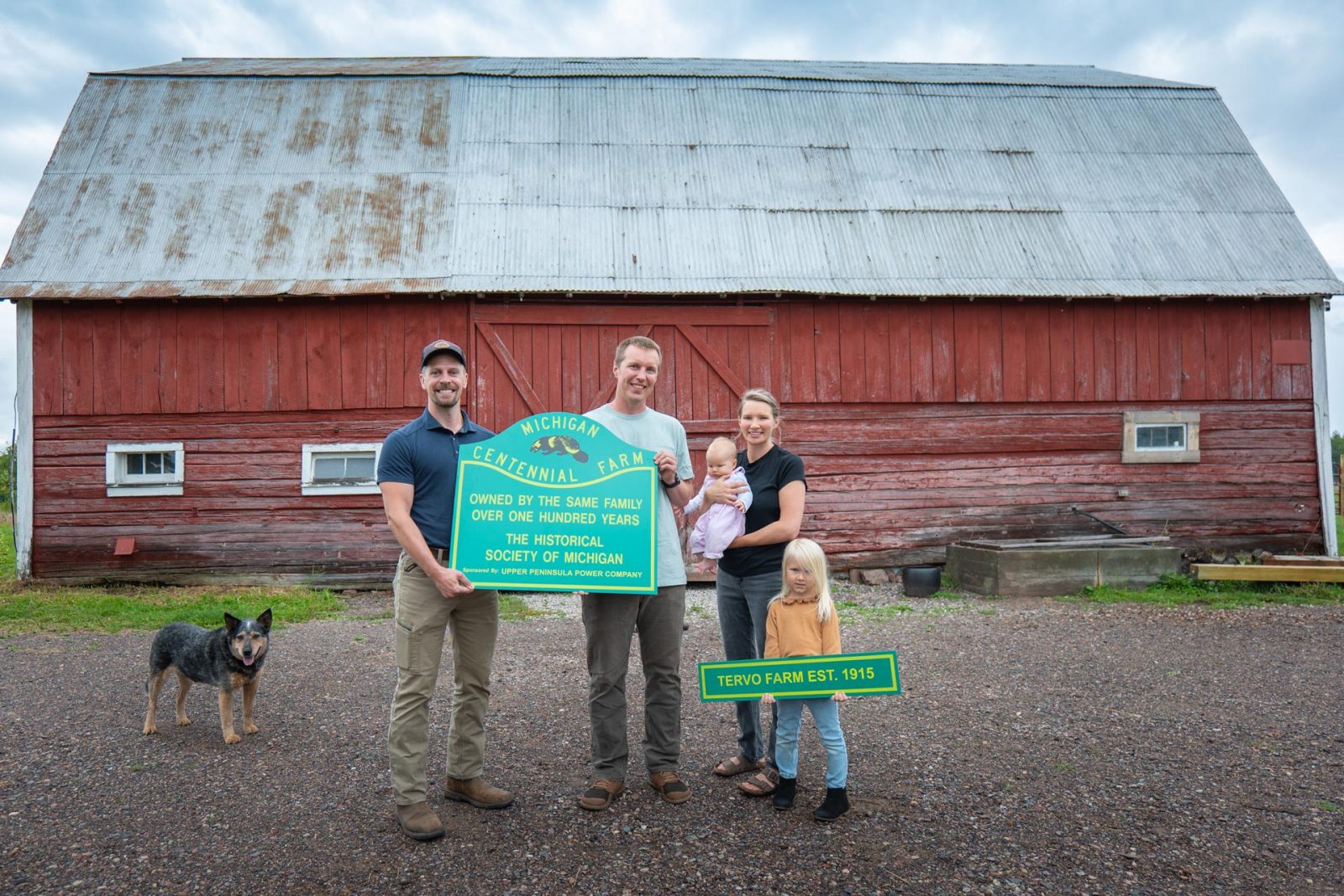An UPPCO representative holds a Centennial Farm sign alongside a family in front of a red barn.