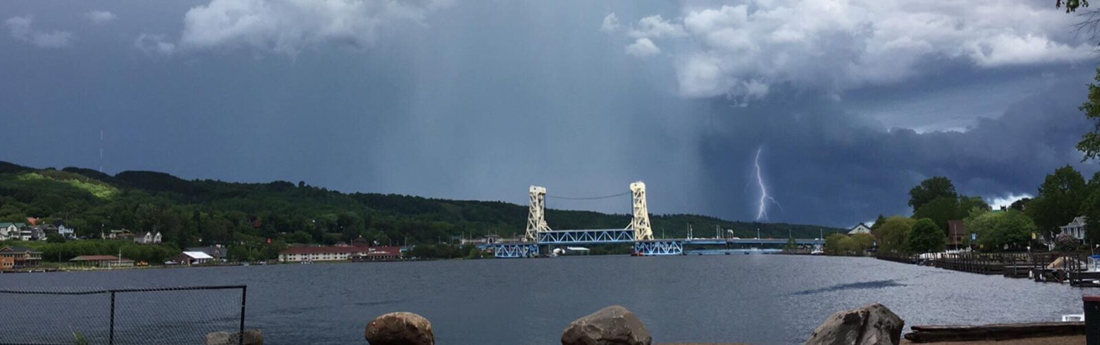 view of water and a bridge with a storm system and visible lightning in the background