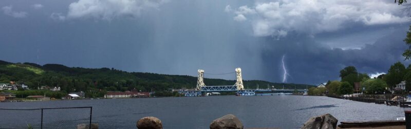 view of water and a bridge with a storm system and visible lightning in the background